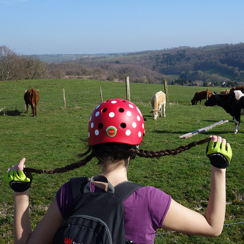 Girl with pig tails wearing a red Melon Bicycle Helmet standing in front of a pasture with cows