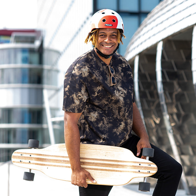 Man holding his Longboard in the city while wearing a colourful Melon Koi Helmet