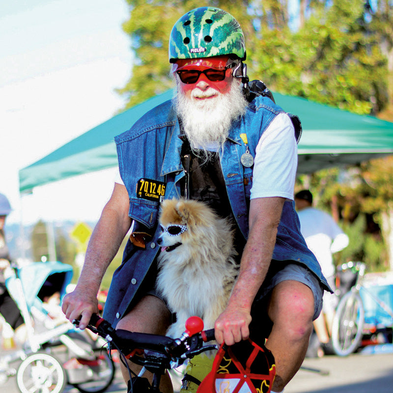 Bearded Man riding a small Bicycle together with his dog while wearing a green Melon Helmet with a red visor