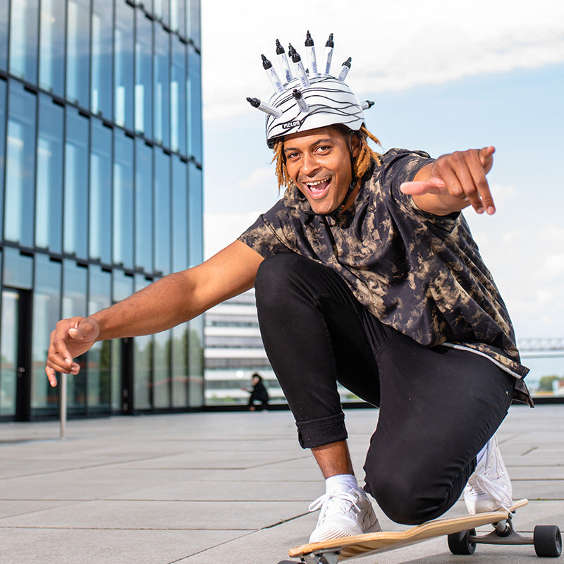 Man kneeling on his Longboard while wearing a white Melon "Scribble" Helmet with sharpies sticking out of the ventilation holes