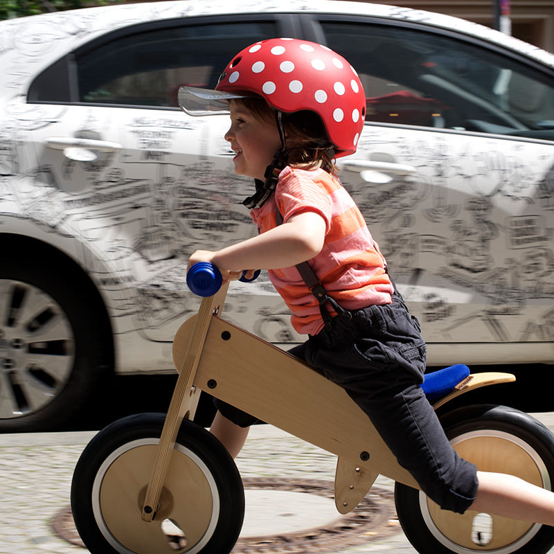 Little girl riding her wooden Bike on the sidewalk wearing a red Melon Bicycle Helmet with a white car in the background that has doodle sketches on it